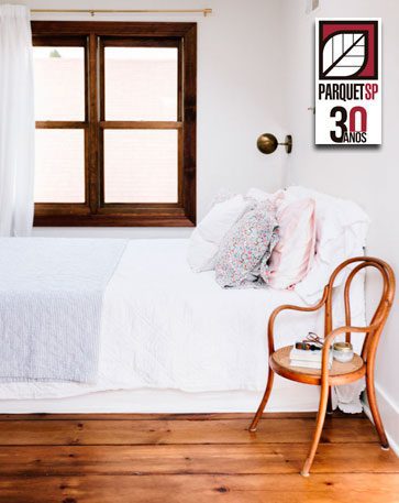 Na foto, um quarto com parede branca e jnaela de madeira marrom escuro. Cama com lençois claros e travesseiros coloridos com cores suaves. Ao lado da cama uma cadeira de madeira marrom médio e piso de madeira da mesma coloração da cadeira.