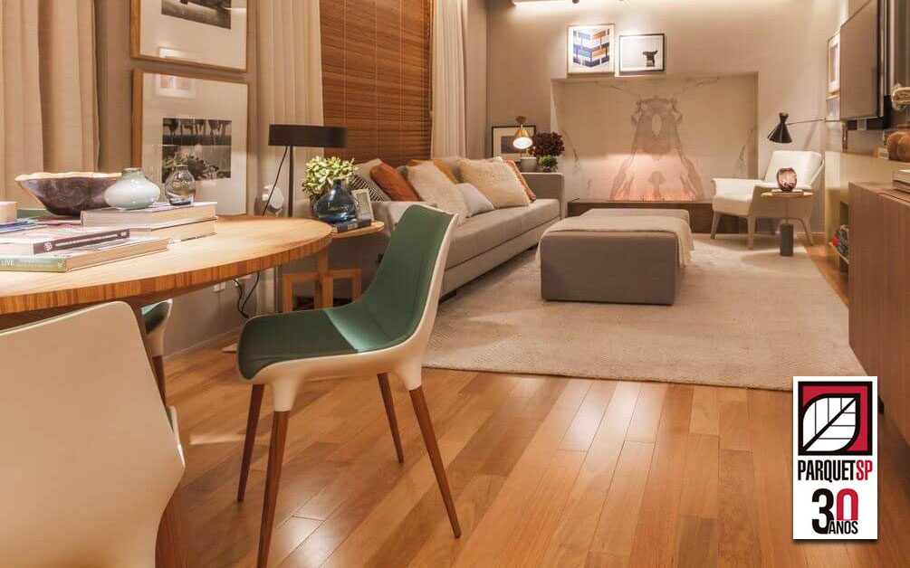 Ao fundo da imagem, há uma sala de estar com sofá, tapete, mesa, cadeira e piso de madeira.