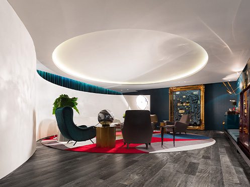 Sala de estar moderna com móveis elegantes, piso escuro, teto com iluminação circular suave, paredes curvas brancas, e uma grande moldura dourada ao fundo.