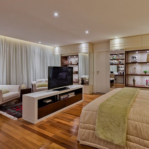 Quarto amplo com piso de madeira, cama de casal, TV em painel central, iluminação embutida e estantes decorativas ao fundo.