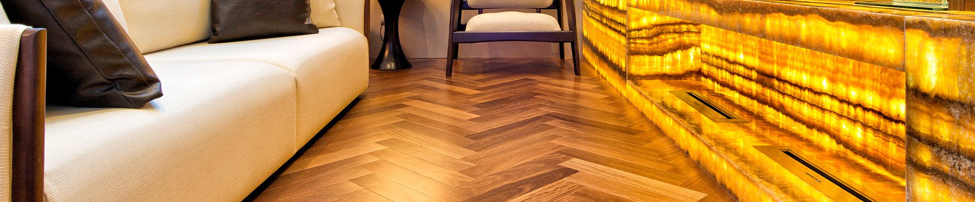 Sala de estar elegante com piso de madeira em padrão espinha de peixe, sofá branco, decoração sofisticada e iluminação suave, criando um ambiente aconchegante e moderno.