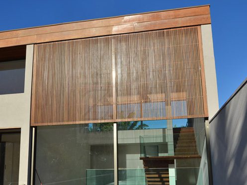 A imagem mostra a fachada de uma casa moderna com brises de madeira verticais cobrindo uma grande janela de vidro, proporcionando estética e funcionalidade.