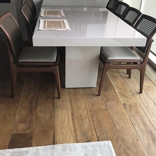 Sala de jantar com mesa branca brilhante e cadeiras de madeira sobre piso de madeira de demolição, criando um ambiente rústico e sofisticado.