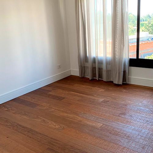 Quarto com piso de madeira rústico e rodapé branco, cortina bege e janela grande com vista para área externa com árvores e construções.