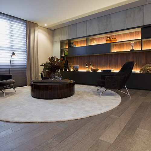 Sala sofisticada com piso de madeira, poltronas escuras, iluminação indireta e decoração moderna.