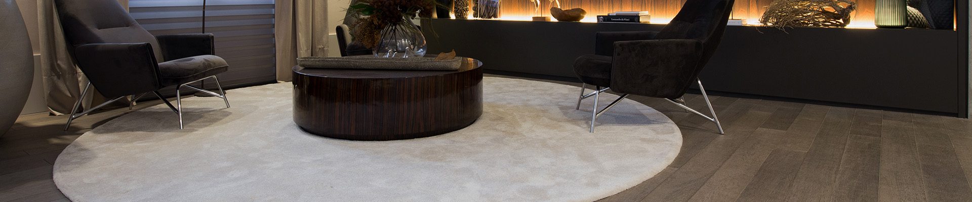 Sala sofisticada com piso de madeira, poltronas escuras, iluminação indireta e decoração moderna.