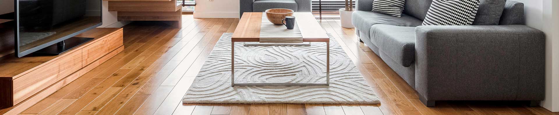 A imagem ilustra uma sala de estar com piso de madeira freijó, mesa de centro, tapere texturizado, sofá cinza com almofadas.