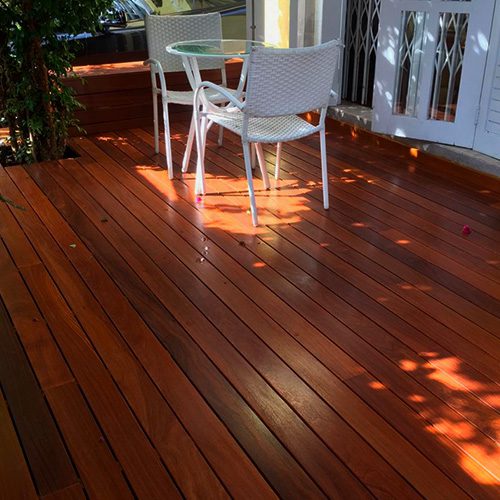 Área externa com deck de madeira, mesa redonda e cadeiras, iluminada por luz natural e cercada por vegetação.