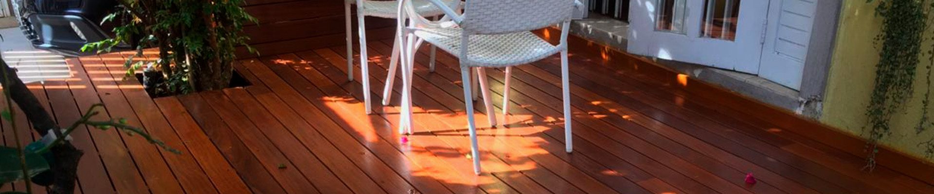 Área externa com deck de madeira, mesa redonda e cadeiras, iluminada por luz natural e cercada por vegetação.