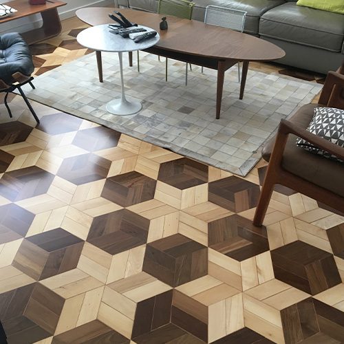 A imagem mostra um piso de madeira escuro com padrão hexagonal e na borda, com uma cor clara, são formadas estrelas com 6 pontas. Há duas cadeiras e uma mesa na sala.