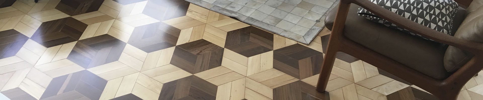 A imagem mostra um piso de madeira escuro com padrão hexagonal e na borda, com uma cor clara, são formadas estrelas com 6 pontas. Há duas cadeiras e uma mesa na sala.