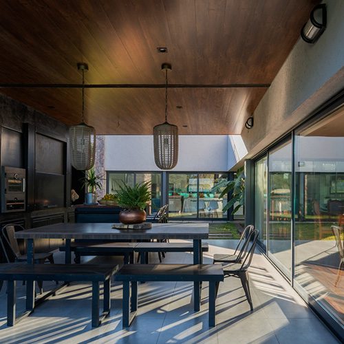 Área gourmet com mesa e bancos, teto em madeira, luminárias pendentes e portas de vidro integrando o ambiente ao espaço externo.