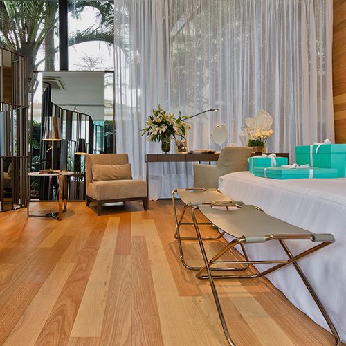 Sala de estar elegante com piso de madeira clara, cortinas brancas, poltronas confortáveis e mesa decorada com flores e presentes azuis.