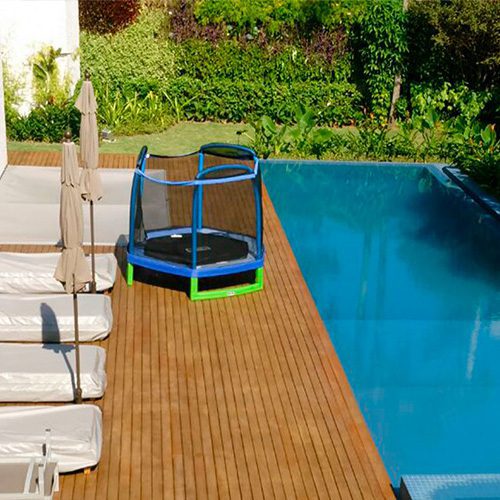 Deck de madeira com espreguiçadeiras ao lado de uma piscina azul, com jardim verde ao fundo e um pula-pula infantil.