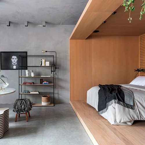 Pessoa deitada em cama com base de madeira clara, quarto moderno com parede cinza e prateleira preta decorada com objetos e livros.