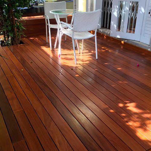Deck de madeira com acabamento impecável em área externa, com mesa e cadeiras brancas sob sombra parcial de uma árvore.