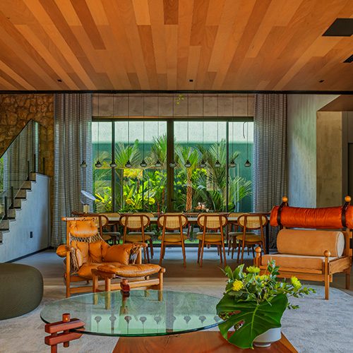Sala de estar moderna com forro de madeira, móveis em tons terrosos e ampla janela com vista para jardim tropical ao fundo.