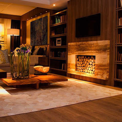 Sala de estar aconchegante com lareira de pedra iluminada, painel de madeira, estante com livros e mesa de centro com arranjo floral.