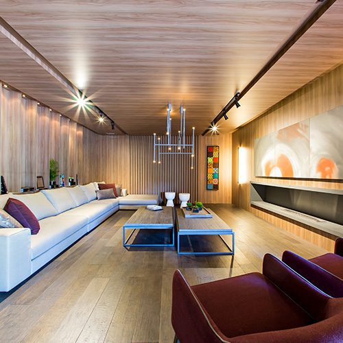 Sala de estar moderna com piso, teto e paredes em madeira, sofá branco em L, poltronas vinho e iluminação aconchegante.