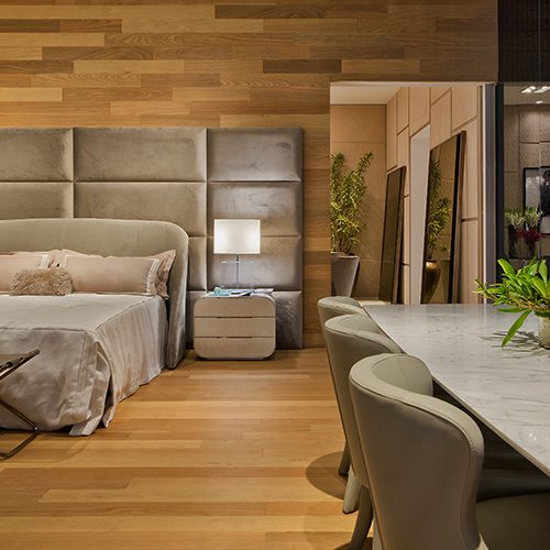 Quarto sofisticado com revestimento de madeira, cabeceira estofada em tons neutros, mesa lateral com abajur e mesa de jantar elegante ao lado.