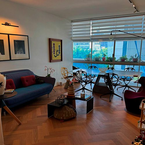 Sala de estar moderna com piso de madeira tauari, sofá azul, cadeiras pretas e janelas amplas que iluminam o ambiente com vista para a varanda.
