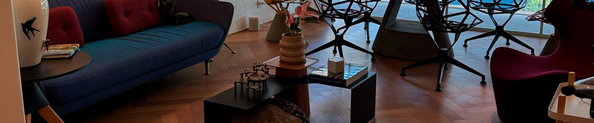Sala de estar moderna com piso de madeira tauari, sofá azul, cadeiras pretas e janelas amplas que iluminam o ambiente com vista para a varanda.