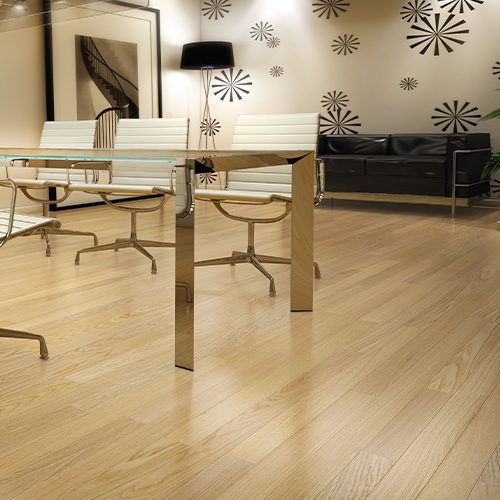 A imagem mostra uma sala de reunião com piso de madeira clara, uma mesa de vidro no centro com 4 cadeiras brancas ao redor e um sofá preto no lado oposto. A parede é decorada com papéis de paredes e um quadro grande.
