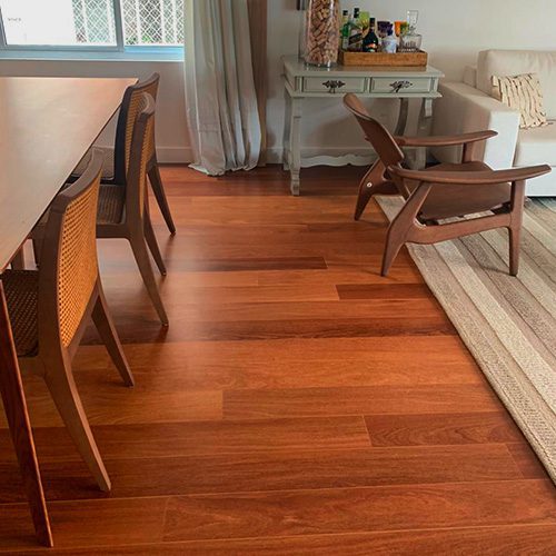 Sala com piso de madeira avermelhada, mesa de jantar, poltronas de design moderno e tapete bege ao lado de sofá claro.