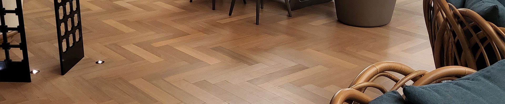 Esta imagem mostra uma sala com um piso feito de tábuas de madeira dispostas em padrão de espinha de peixe. Há uma mesa e cadeiras no centro da sala e uma grande janela.