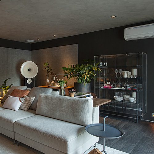 Sala moderna com piso de madeira escura, sofá claro, decoração sofisticada, plantas e prateleira de vidro com utensílios e objetos decorativos.