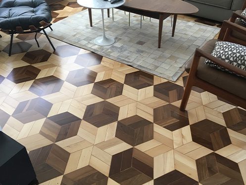 Piso de madeira com padrão geométrico em tons variados, criando um efeito tridimensional, em uma sala de estar com móveis modernos e tapete neutro.