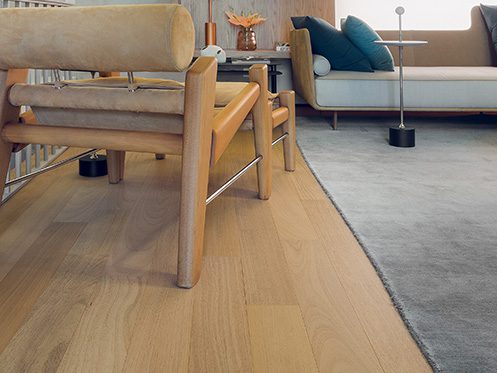Sala de estar moderna com piso de madeira clara, poltronas de madeira e tecido bege, sofá cinza com almofadas, e mesa lateral com decoração minimalista.