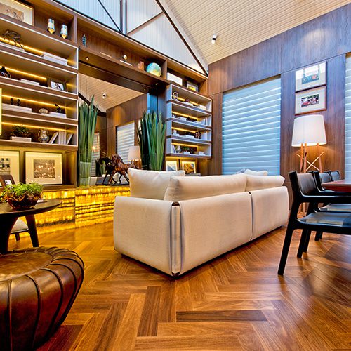 Sala de estar elegante com piso de madeira em padrão espinha de peixe, sofá bege, prateleiras iluminadas com objetos decorativos e uma atmosfera aconchegante.