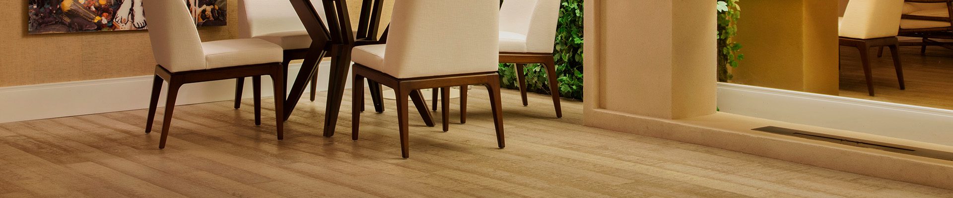 Sala de jantar com piso de madeira, mesa com cadeiras estofadas, parede coberta por plantas verdes, luminária de piso e quadro decorativo.