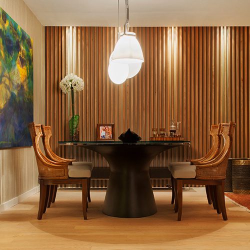 Sala de jantar com painel ripado de madeira na parede, piso de madeira clara, mesa redonda, cadeiras estofadas, luminária pendente, quadro decorativo e objetos de decoração.