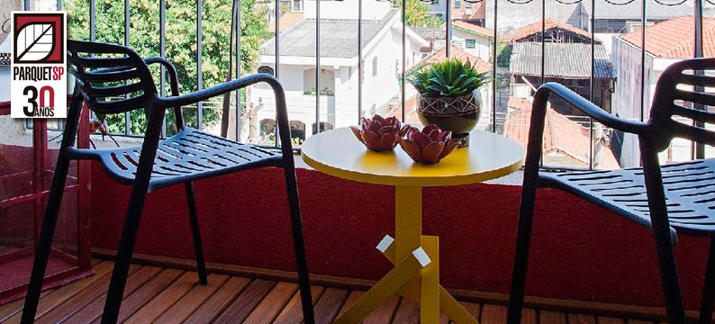 Sacada de apartamento com deck de madeira, mesa de centro amarela e duas cadeiras pretas