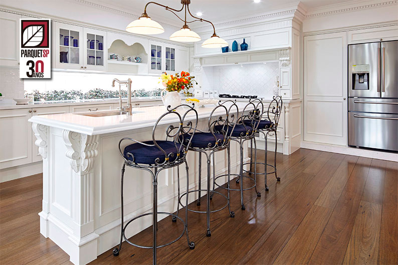 Cozinha americana com bancada branca e cadeiras altas de ferro. No chão, piso de madeira.