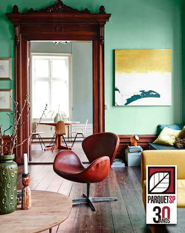 Na foto, uma sala com piso de madeira escuro, parede verde claro e batente da porta da mesma cor do piso. No canto um sofá da cor bege e ao centro, uma poltrona aparentemente de couro na cor vermelho queimado