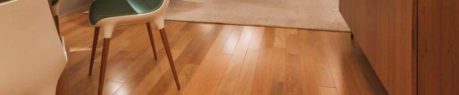Ao fundo da imagem, há uma sala de estar com sofá, tapete, mesa, cadeira e piso de madeira.